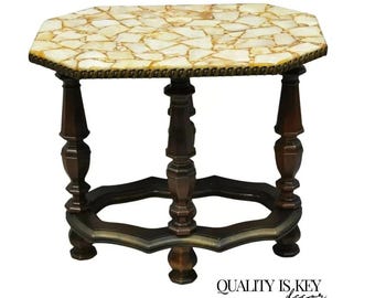 May include: Octagonal side table with a unique, mosaic-like top made of small, irregular pieces of white and gold stone. The table has a dark wood base with intricate carvings and a decorative brass trim around the top.