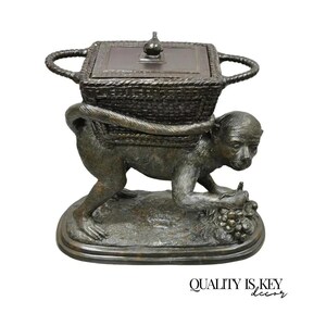 May include: A bronze-colored monkey sculpture carrying a basket on its back. The monkey is depicted in a crouching position, holding grapes. The basket has a lid and handles. The base is oval-shaped.