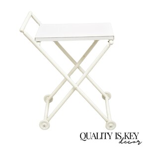 May include: White folding serving cart with a white top and wheels. The cart is made of metal and has a criss-cross design.