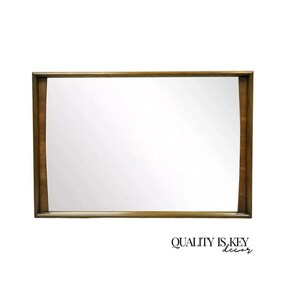 May include: A large, rectangular mirror with a wood frame. The frame has a curved design on the top and bottom edges. The mirror is reflecting a white wall.
