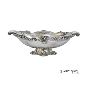 May include: A silver-colored, ornate serving bowl with a scalloped rim and decorative floral accents. The bowl sits on a pedestal base and features intricate detailing. The words "QUALITY IS KEY decor" are printed in black at the bottom right.