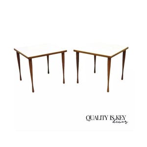 May include: A pair of mid-century modern end tables with white laminate tops and brown wooden legs. The tables have a simple, elegant design.