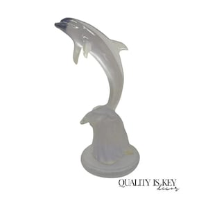 May include: A clear, frosted glass dolphin figurine in a leaping pose. The dolphin is mounted on a round base, with the text "QUALITY IS KEY decor" in the bottom right corner. The figurine is a decorative item.