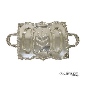 May include: A silver-colored rectangular serving tray with ornate handles and a decorative floral design in the center. The tray has three compartments and is made of a shiny metal. The text "QUALITY IS KEY decor" is in the bottom right corner.