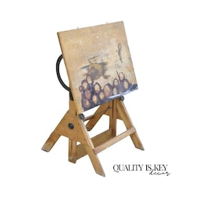 May include: A vintage wooden drafting table with a tilting surface. The table has a natural wood finish, a black adjustment arm, and a sturdy, angled base. The surface shows signs of use, with paint marks and wear.