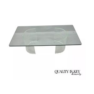 May include: A clear glass coffee table with a rectangular top and two clear acrylic bases. The bases are shaped like a curved 'C' and are joined together by the glass top.