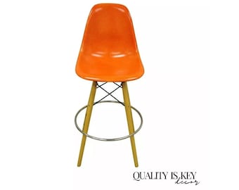 May include: Orange fiberglass bar stool with a wooden base and a chrome footrest. The stool has a simple, modern design.