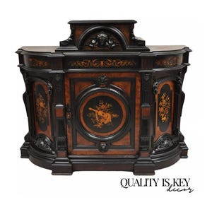 May include: A large, antique, dark wood cabinet with intricate carvings and marquetry. The cabinet has two doors with a central panel featuring a floral design and a musical instrument. The cabinet is in excellent condition and would be a beautiful addition to any home.