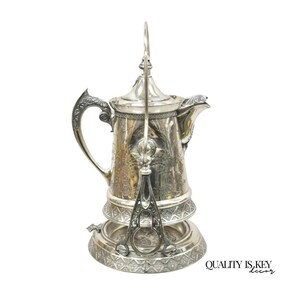 May include: A silver-toned metal teapot with an ornate handle, a hinged lid, and a decorative spout. The pot rests on a matching circular base with a textured geometric pattern. The text QUALITY IS KEY decor is visible in the bottom right corner.