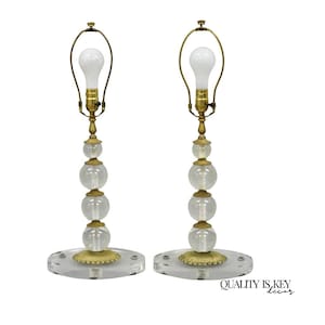 May include: Pair of vintage table lamps with clear glass orb accents. Each lamp features a brass-toned base and stem, topped with a white light bulb. The lamps sit on a clear, oval-shaped base. The lamps are a decorative lighting fixture.