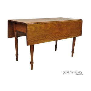 May include: A wooden drop-leaf table with a warm, medium-brown finish. The table has a rectangular top with two drop leaves and four turned legs. The table is designed to save space when the leaves are down. The text "QUALITY IS KEY decor" is visible.