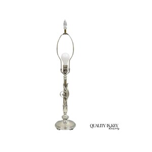 May include: A silver-toned table lamp with a decorative, ornate design. The lamp features a bulb and a clear, pointed finial at the top. The base is round and the stem has a swirling, floral-like pattern. The text "QUALITY IS KEY decor" is visible.
