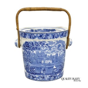 May include: A blue and white ceramic ice bucket with a landscape design. The bucket has a woven handle and small wooden side handles. The design features a scenic view with trees, buildings, and a sailboat. The text "QUALITY IS KEY decor" is visible.