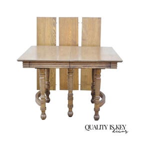 May include: A light brown, rectangular dining table with ornate, turned legs. The table has a multi-layered edge and includes three additional leaves. The text "QUALITY IS KEY decor" is visible in the lower right corner.