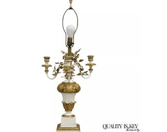 May include: A white and gold table lamp with a candelabra design. The lamp features a white base with gold accents, a decorative floral design, and two candle holders. The lamp has a light bulb and a lampshade holder.