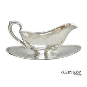 May include: A silver gravy boat with a matching oval-shaped saucer. The gravy boat has a curved handle and a spout for easy pouring. The set is made of a shiny, reflective metal, and the words "QUALITY IS KEY decor" are printed on the bottom right.
