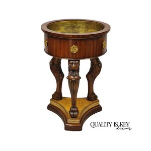 May include: A brown wooden pedestal table with a round top and ornate lion's paw feet. The top has a gold-colored interior. Gold-colored decorative accents are on the side of the table. The base is a light brown color with a gold trim.
