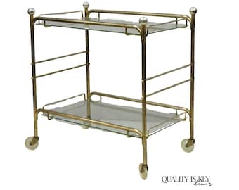 May include: A gold-coloured, two-tiered bar cart with clear glass shelves. The cart has a rectangular shape with rounded corners and small wheels for easy movement. The frame is made of metal and has a vintage aesthetic.