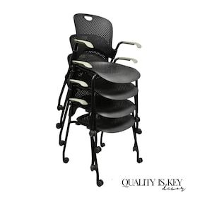 May include: Stack of four black office chairs with mesh backs and gray armrests. The chairs are stacked on top of each other with the legs of the bottom chair visible.