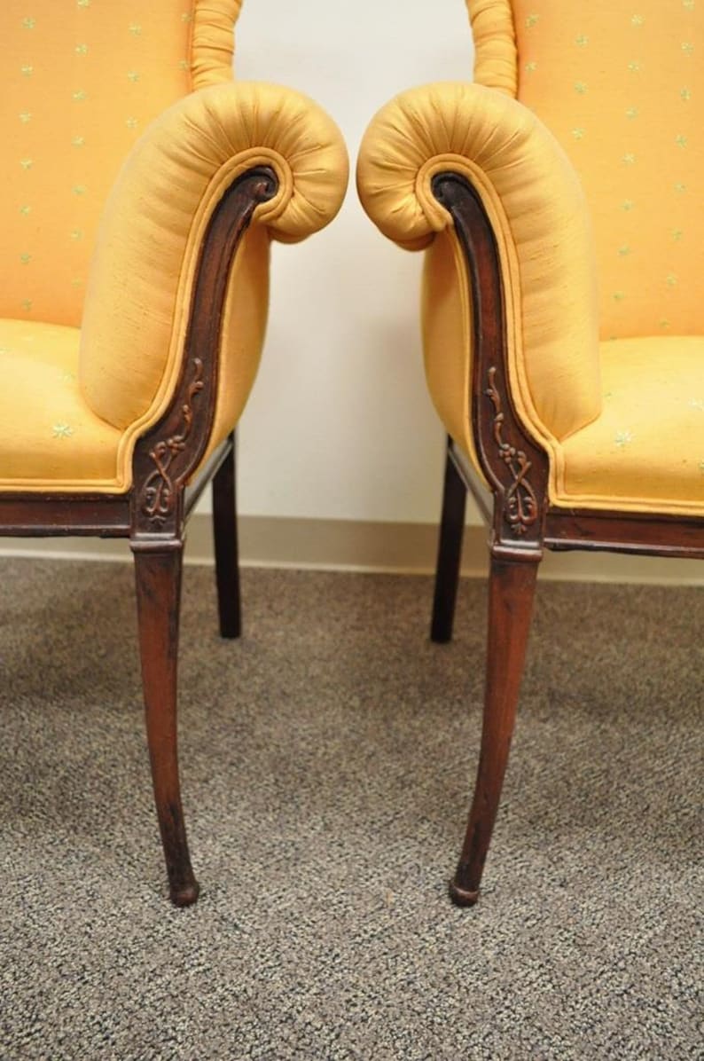 May include: A pair of yellow upholstered chairs with wooden frames and carved details. The chairs have a scrolled back and a curved armrest.