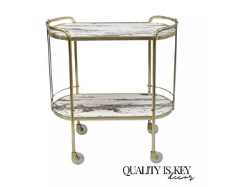 May include: A gold metal serving cart with two oval-shaped shelves. The shelves are made of glass with a white and brown wood-grain pattern. The cart has four wheels.
