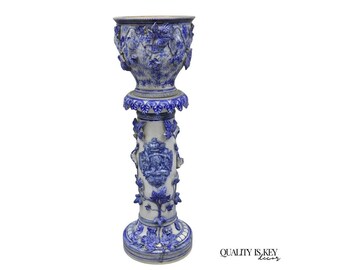 May include: A tall ceramic pedestal planter featuring a white base with intricate blue relief carvings of grapevines and leaves. The cylindrical column supports a wide, matching bowl-shaped planter. The text QUALITY IS KEY decor is visible.