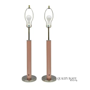 May include: Two matching table lamps with a mid-century modern design. Each lamp features a cylindrical, rose-gold colored base, a slender brass stem, and a white lightbulb. The lamps are set on a silver-colored circular base.