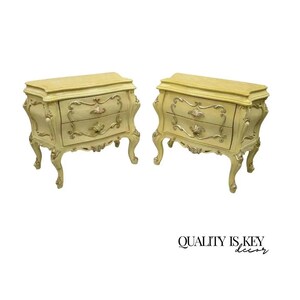 May include: Two antique cream-colored nightstands with ornate detailing. Each nightstand features two drawers with gold-colored hardware and decorative carvings. The curved legs and edges add to the vintage aesthetic. The tops have a light yellow finish.