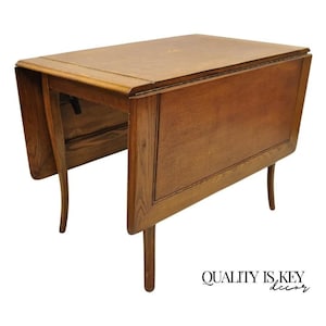 May include: A vintage wooden drop leaf table with a light brown finish. The table has a rectangular top with rounded corners and four legs. The table is in a folded position, with one leaf down and the other leaf up.