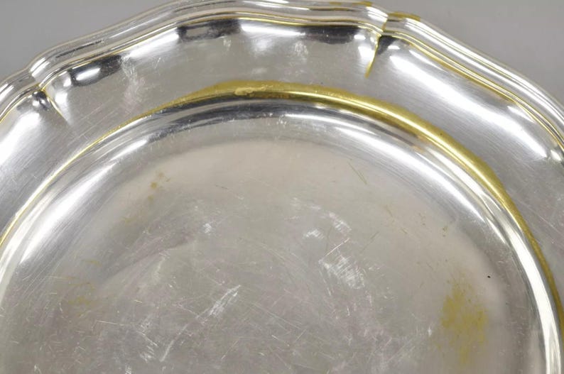 Puede incluir: Un plato de servir de plata con un borde dorado. El plato tiene un borde festoneado y una superficie lisa y reflectante.