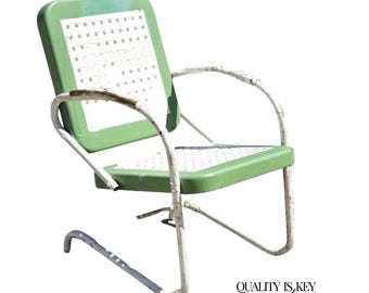 May include: Vintage green and white metal patio chair with a perforated seat and backrest. The chair has a curved, white metal frame and armrests, showing signs of wear. The chair is designed for outdoor use.