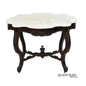 May include: An antique, dark wood side table with a white marble top. The table has ornate carvings on the apron and legs. The legs are curved and end in small feet. The table is a classic piece of furniture.