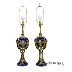 May include: Pair of antique table lamps with a dark blue ceramic base and ornate brass accents. The lamps feature a bulb, a brass neck, and a decorative design around the base. The lamps are on a white background.