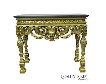 May include: A gold-painted console table with a black marble top. The table features intricate carvings and a decorative design with a central face.