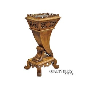 May include: A tall, light brown wooden plant stand featuring an intricate, open-work carved rectangular top section with geometric patterns. The base is supported by a curved, pedestal-style column and four ornate, scrolled feet. Text reads: QUALITY IS KEY decor.