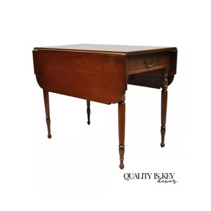 May include: A cherry-stained, drop-leaf table with a single drawer. The table has four turned legs and a rectangular top with rounded edges when the leaves are down. The drawer has a brass pull. The table is approximately 30 inches tall.