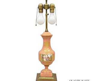 May include: A vintage table lamp with a peach-colored ceramic base and brass accents. The lamp features a double bulb fixture and a decorative panel with a painted scene. The base is adorned with gold floral designs and rests on a square, ornate base.