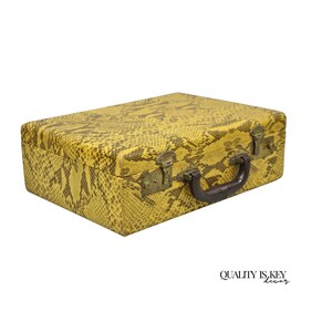 May include: A rectangular yellow suitcase with a snakeskin pattern. The suitcase has a dark brown handle and brass-colored clasps. The text "QUALITY IS KEY decor" is in the bottom right corner.