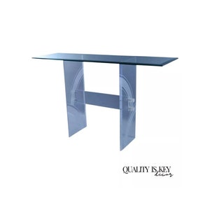 May include: A clear acrylic console table with a glass top and a geometric design on the legs. The table has a lower shelf.