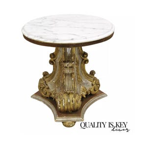 May include: A round marble top table with a gold and white painted base. The base is ornately carved with scrolls and floral details. The table is on a white background.
