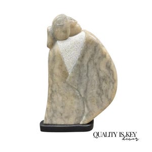 May include: A stone sculpture of a person in a flowing robe, with the head tilted upwards. The sculpture has a light beige and gray color palette, with a textured white area on the chest. It is mounted on a black base. The text "QUALITY IS KEY decor" is in the bottom right corner.