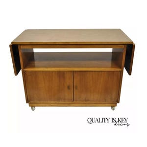 May include: A mid-century modern wooden bar cart with two doors, a shelf, and drop-down leaves. The cart has a light brown wood finish and is on wheels.