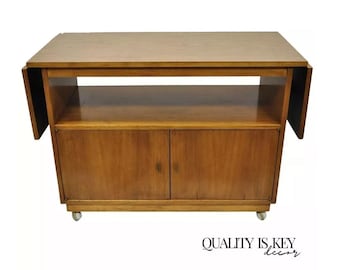 May include: A mid-century modern wooden bar cart with two doors, a shelf, and drop-down leaves. The cart has a light brown wood finish and is on wheels.