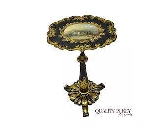 May include: A black and gold painted side table with a round top and a single pedestal base. The top features a painted scene of a building and trees. The base has a decorative, scalloped design.