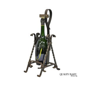 May include: A dark green wine bottle is held in a wrought iron wine bottle holder. The holder has a decorative design with twisted metal supports and a handle. The bottle is partially visible, with a yellow label. The holder is on a white background.