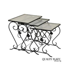 May include: A set of three nesting tables with black metal frames featuring scrollwork and leaf accents. Each table has a square top with a textured, light-colored surface. The text "QUALITY IS KEY decor" is printed in the bottom right corner.