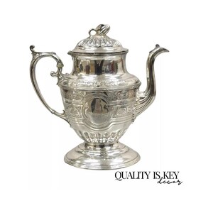 May include: A silver teapot with an ornate design and a round body. The teapot has a lid with a knob on top and a spout on the side. The teapot is sitting on a white background.