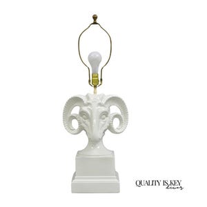 May include: White ceramic table lamp featuring a ram's head design. The lamp has a square base and a brass-colored lamp holder. The lamp includes a light bulb. The text "QUALITY IS KEY decor" is visible.