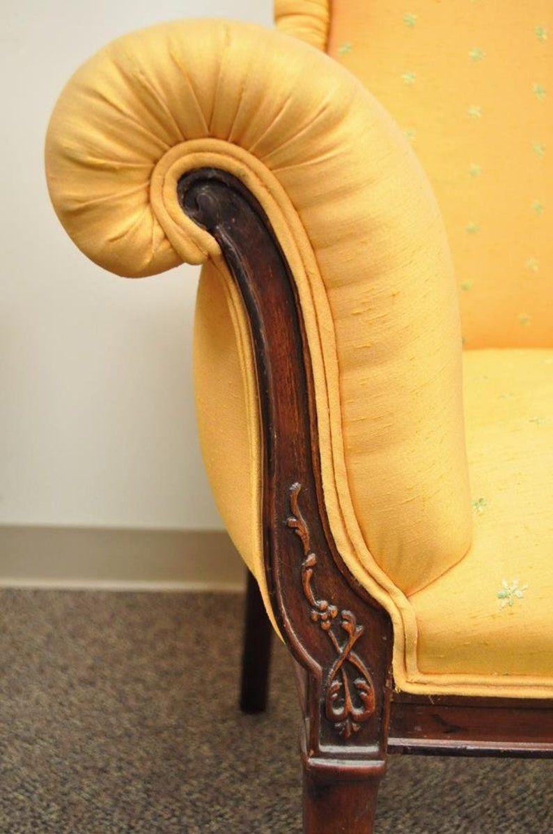 May include: A yellow fabric armchair with a carved wooden armrest and a scrolled design. The chair has a tufted back and a rolled armrest.