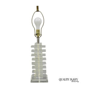May include: A clear acrylic table lamp with a stacked rectangular base. The lamp has a brass-colored neck and a white light bulb. The lamp is a modern design and is suitable for a living room or bedroom.